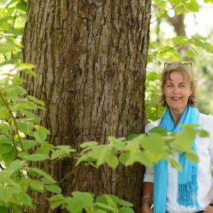 Een vrouw staat naast een dikke boom in het park. Het is voorjaar en het licht schijnt zacht door het prille groen. Ze kijkt blij en tevreden.