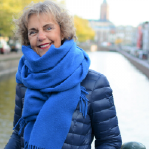 Vrouw met een blauwe jas en een dikke blauwe sjaal staat op de brug over een gracht in Den Haag. In de verte is de toren van een kerk zichtbaar.