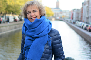 Vrouw met een blauwe jas en een dikke blauwe sjaal staat op de brug over een gracht in Den Haag. In de verte is de toren van een kerk zichtbaar.