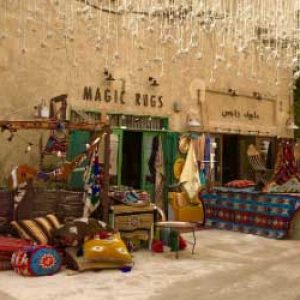 Een winkel met handgemaakte tapijten in Dubai. Voor de winkel liggen talloze kleurige tapijten en kussens. De winkel draagt de naam Magic Rugs, magische tapijten en. De foto ademt een Oosterse sfeer.