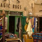 Een winkel waar met de handtapijten worden gemaakt in Dubai. Voor de winkel een uitstalling van allerlei kleurige tapijten. Op de muur van de winkel staat de naam Magic Rugs, magische tapijten. De foto is kleurig en ademt een oosterse atmosfeer.i.