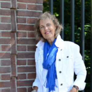 Een vrouw met een wit jasje en blauwe sjaal staat bij een muur. Ze glimlacht.