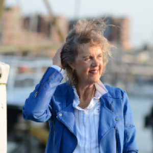 Een vrouw staat aan de haven. Haar haar waait omhoog en zij houdt het in bedwang met haar rechterhand. Zij draagt een felblauw jasje en een blauw wit gestreepte blouse. Zij kijkt in de verte en glimlacht.