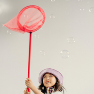 Een jong meisje met donker haar en een roze hoedje op haar hoofd, probeert met een oranje vlindernet zeep bellen te vangen.