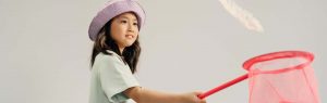 Een jong meisje met donker haar en een roze hoedje op haar hoofd, Probeert met een oranje vlindernet een wit veertje te vangen.