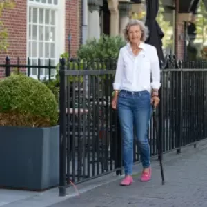 Een vrouw van middelbare leeftijd loopt met een wandelstok over de Kneuterdijk in den Haag. Ze loop langs het hek van een prachtig oud pand. Het is mooi weer en zij draagt een witte blouse en blauwe jeans. Zij kijkt lachend in de lens.